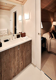 Salle de bains d'une chambre du chalet. Sol en pierre, grand miroir au dessus du meuble lavabo avec plan de travail en pierre et porte en chêne.