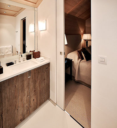 Salle de bains d'une chambre du chalet. Sol en pierre, grand miroir au dessus du meuble lavabo avec plan de travail en pierre et porte en chêne.